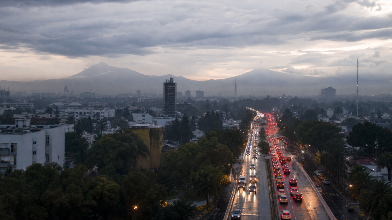 Tráfico vehicular en una avenida de la Ciudad de México al amanecer, con pavimento mojado y cielo nublado.